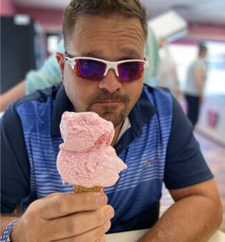 Adult man wearing sunglasses, sitting outside eating an ice cream cone with 2 scoops of pink ice cream